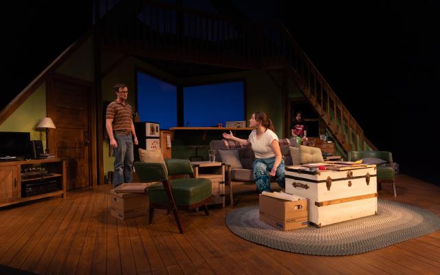 Living room set of a play with a male actor standing on the left talking to a female actor seated on the couch, both are in intese conversation.