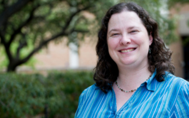 A white woman with wavy shoulder-length brown hair, freckles, and a blue and white shirt is smiling outside of University Hall. An oak tree and a hedge are in the background.