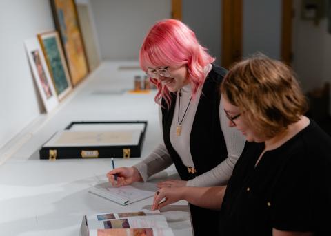 Two University of Lethbridge museum studies intern students smiling while looking through an art textbook and writing notes. 