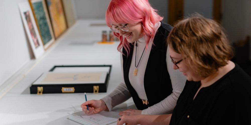 Two University of Lethbridge museum studies intern students smiling while looking through an art textbook and writing notes. 