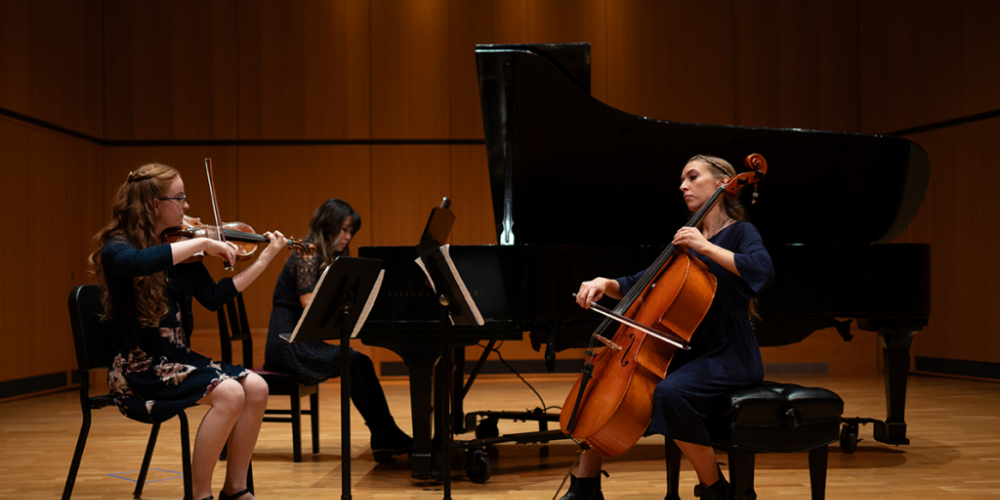 Left to right on recital hall stage (light wood floors & wood wall paneling): Alayna McNeil sitting playing violin, Anna Jeong playing on black grand piano, Brenna Le May, sitting playing cello.