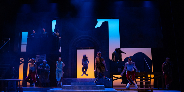 A scene from the University of Lethbridge's production of Jesus Christ Superstar. A number of actors hovering in the air mid-dance. Set is cast in a darker blue light. Backdrop is backlit with lighter blue, magenta and yellow.