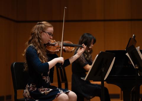 Alayna McNeil is seated playing the violin and Anna Jeong is seated playing the black grand piano.